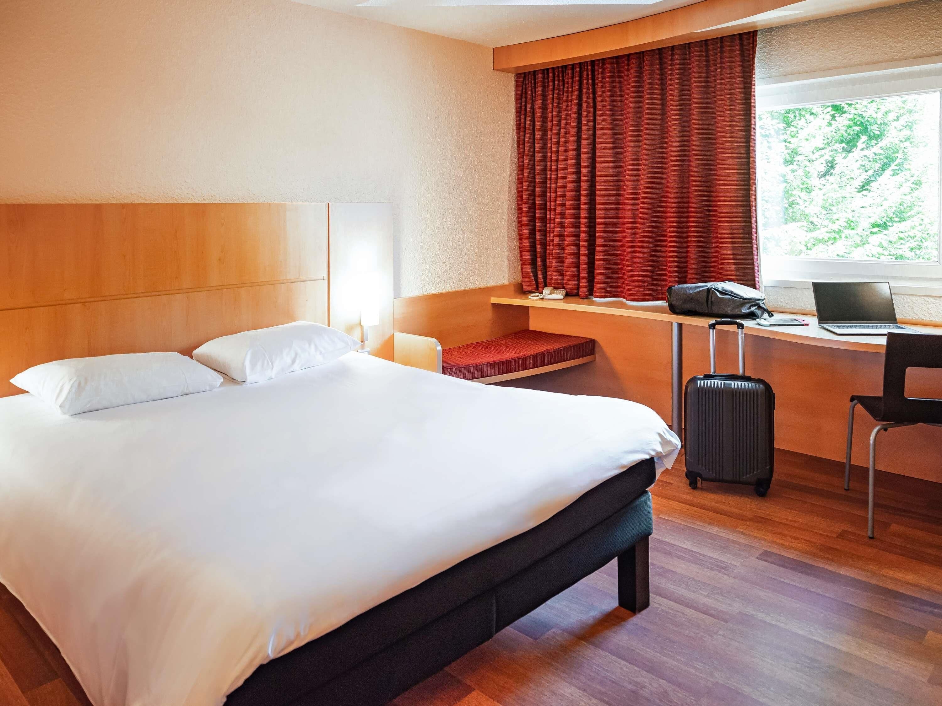 Een hotelkamer met een bed, een bureau met een laptop, een koffer en een raam met gordijnen.
