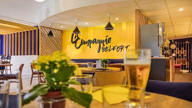 A modern restaurant interior with a curved ceiling, pendant lighting, and a prominent wall sign reading "Compagnie BELFORT".