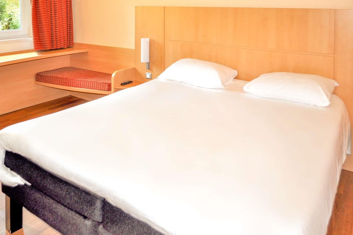 Ein Hotelzimmer mit Bett, Nachttisch, Lampe und einem Fenster mit Vorhängen.