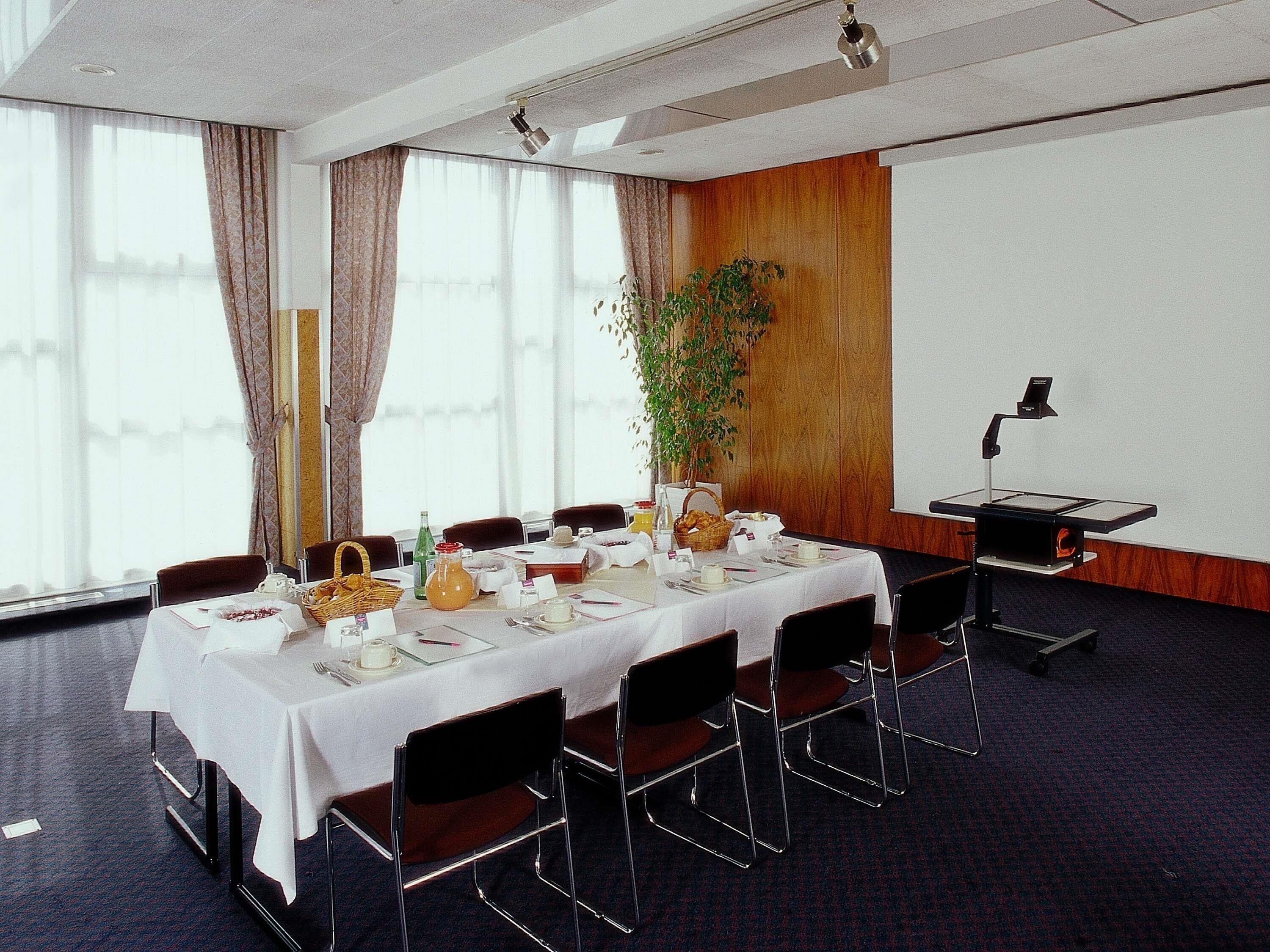 Un salón de conferencias con una larga mesa, sillas, un proyector y una planta.