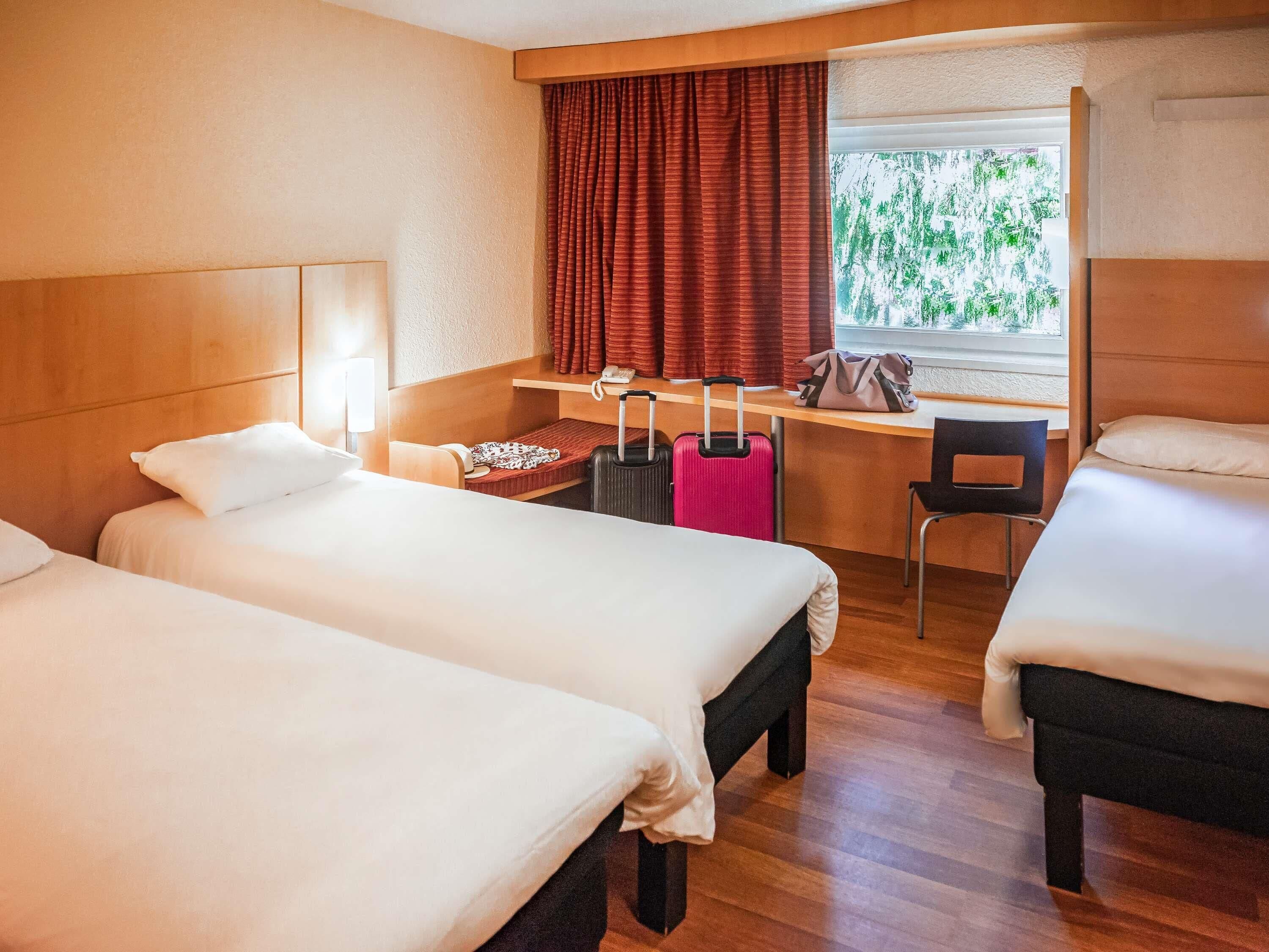 Habitación de hotel con dos camas, un escritorio, una silla, una maleta y una ventana con vistas a árboles.
