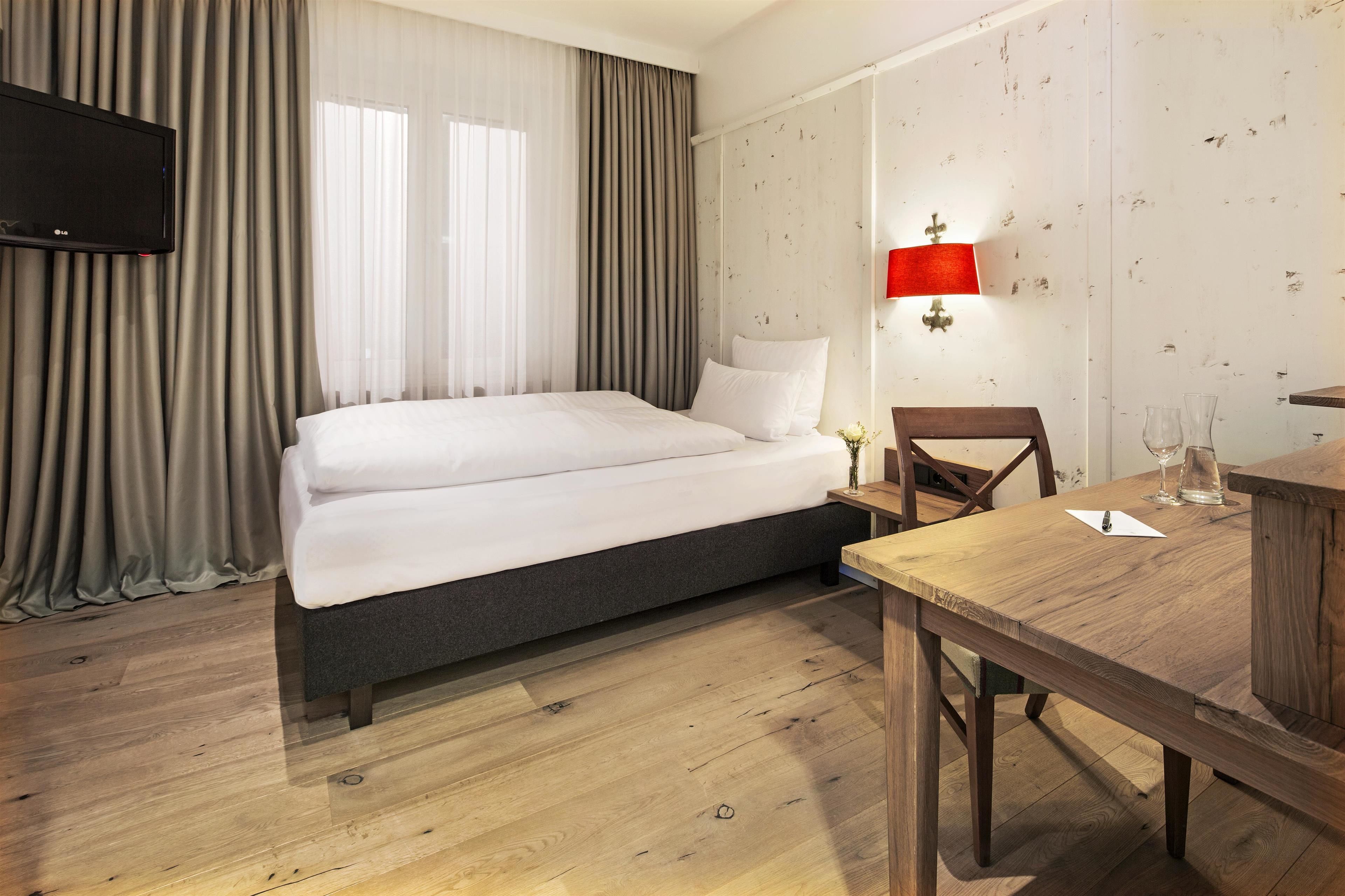 Una habitación de hotel con cama, una mesa de madera, una silla, un televisor y una lámpara de pared.