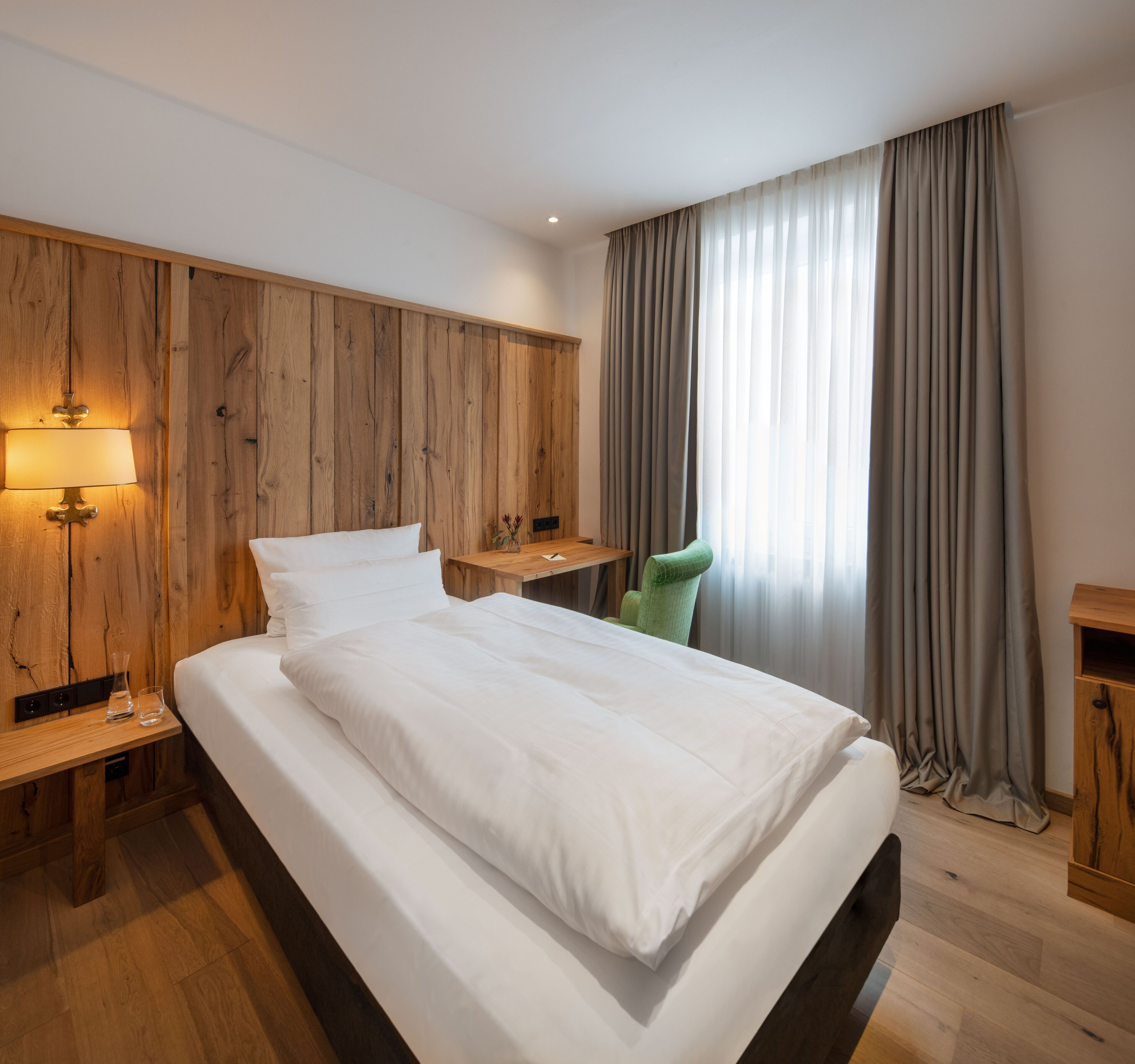 A modern hotel room with a wooden headboard, a large bed with white linens, a bedside table with a lamp, a green chair, and a window with grey curtains.