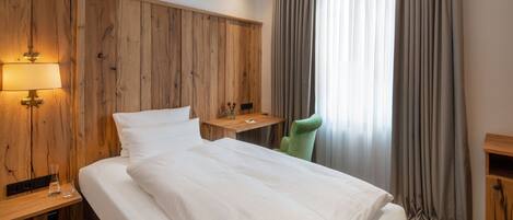 Une chambre d’hôtel moderne avec une tête de lit en bois, un grand lit avec du linge de lit blanc, une table de chevet avec une lampe, un fauteuil vert et une fenêtre avec des rideaux gris.