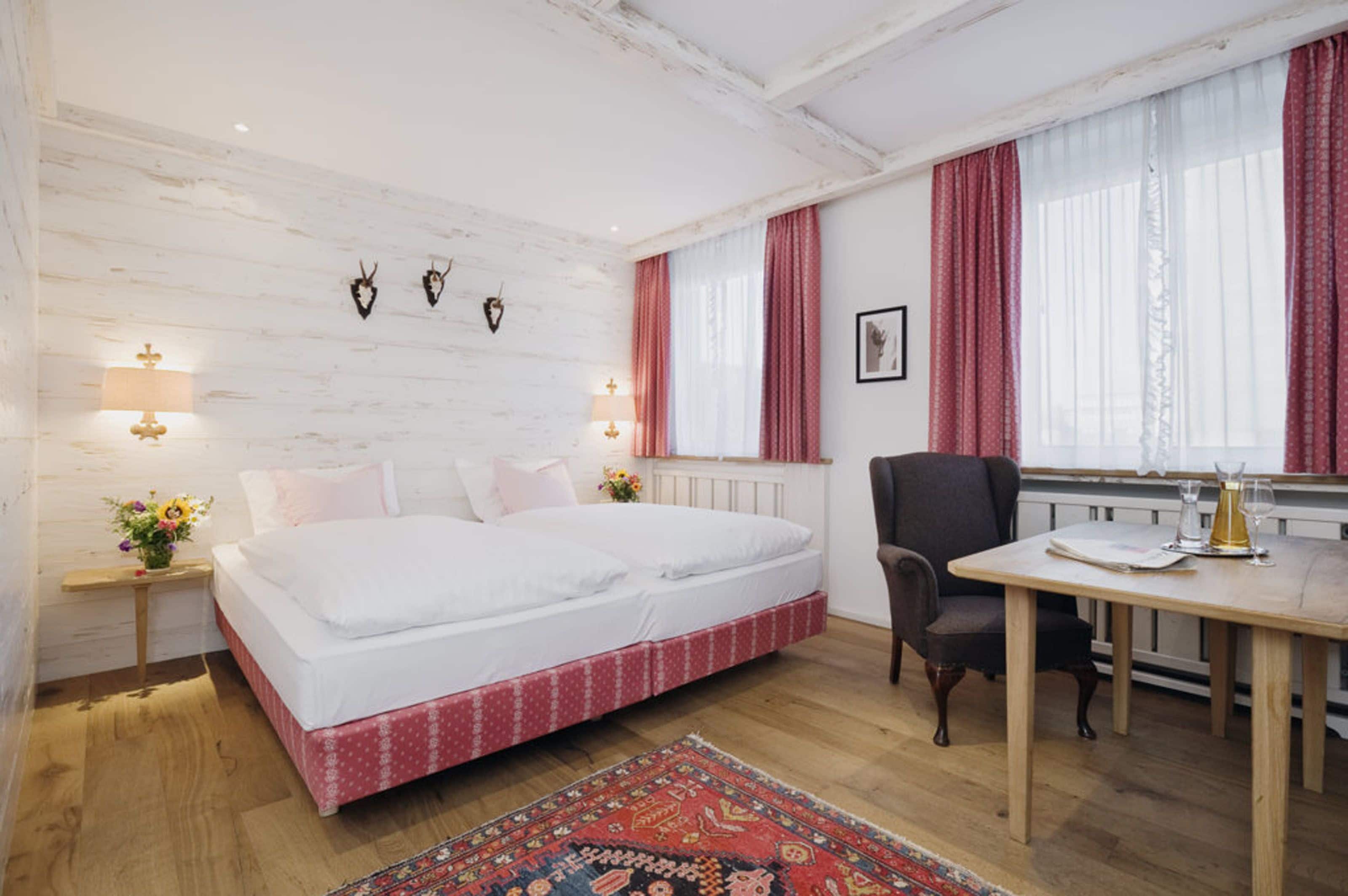 Un dormitorio con cama, mesitas de noche, una silla, una mesa y una ventana con cortinas.