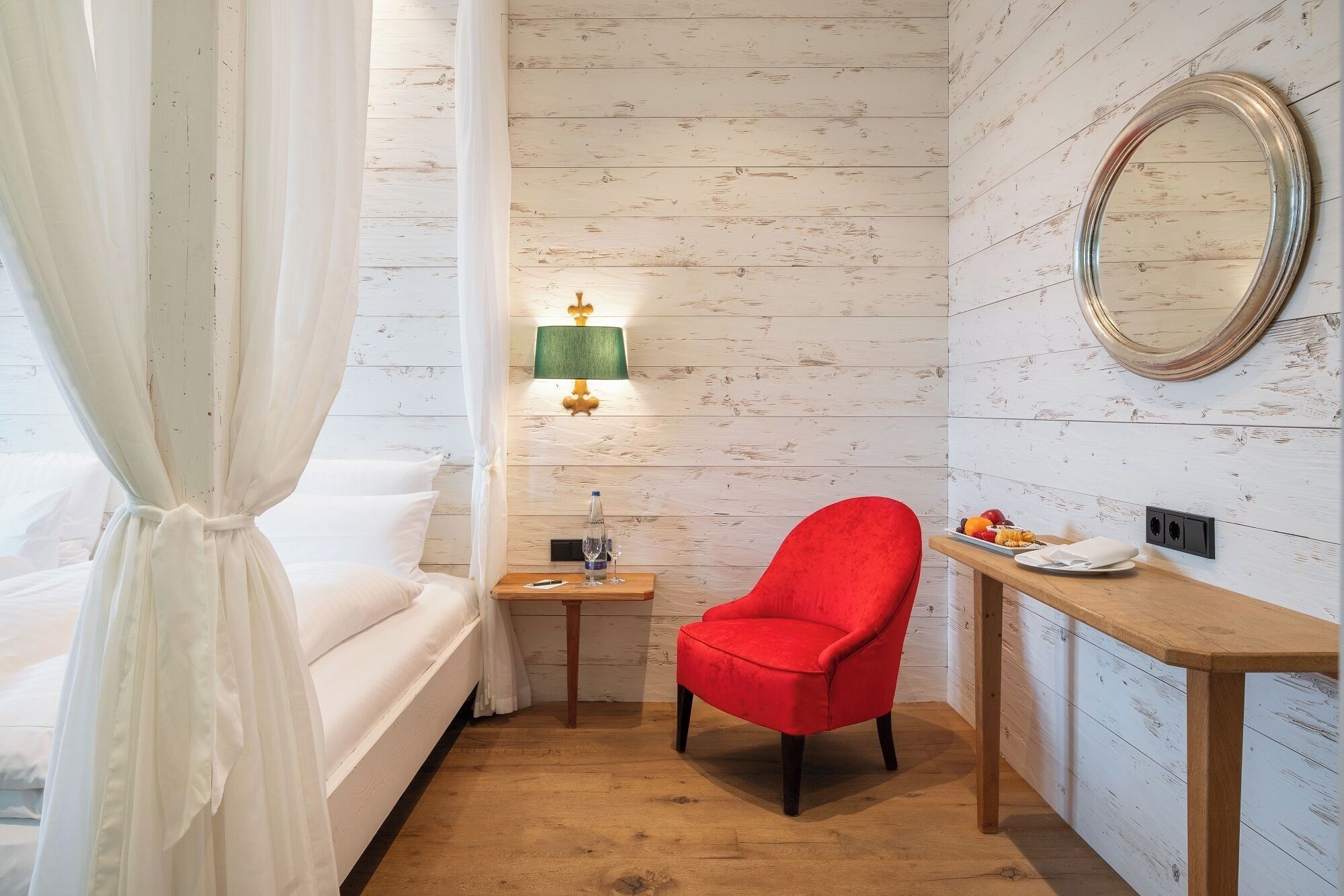 A hotel room with a bed, a red chair, a small table with a lamp, and a mirror on the wall.