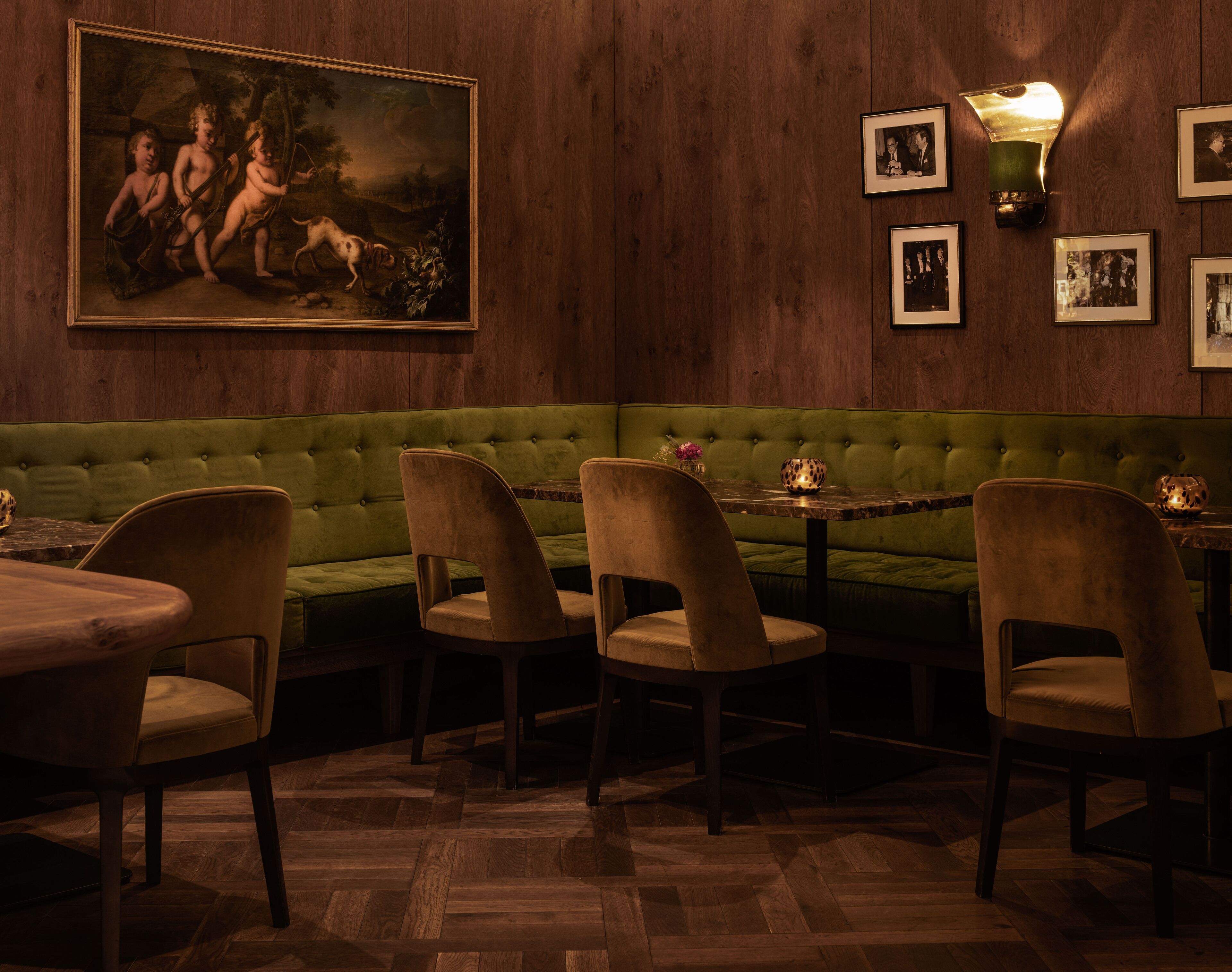 Un restaurante con una banqueta tapizada de verde, mesas y sillas de madera. Hay un cuadro enmarcado en la pared y una lámpara de pared.