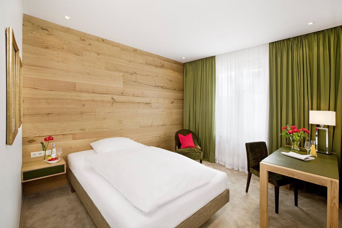 Een hotelkamer met een houten hoofdeinde, een bed met witte lakens, een groene stoel, een klein tafeltje met een lamp en bloemen, en een groen gordijn.