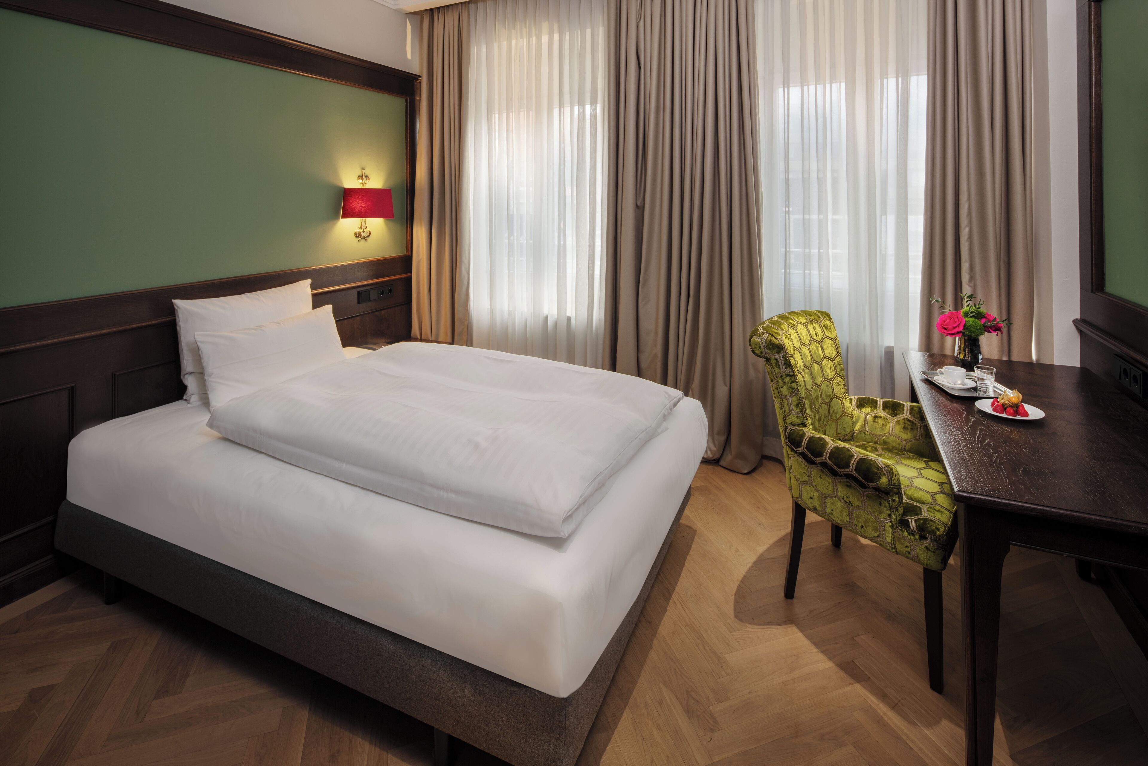 A hotel room with a large bed, a desk with a chair, a lamp, and a window with curtains.