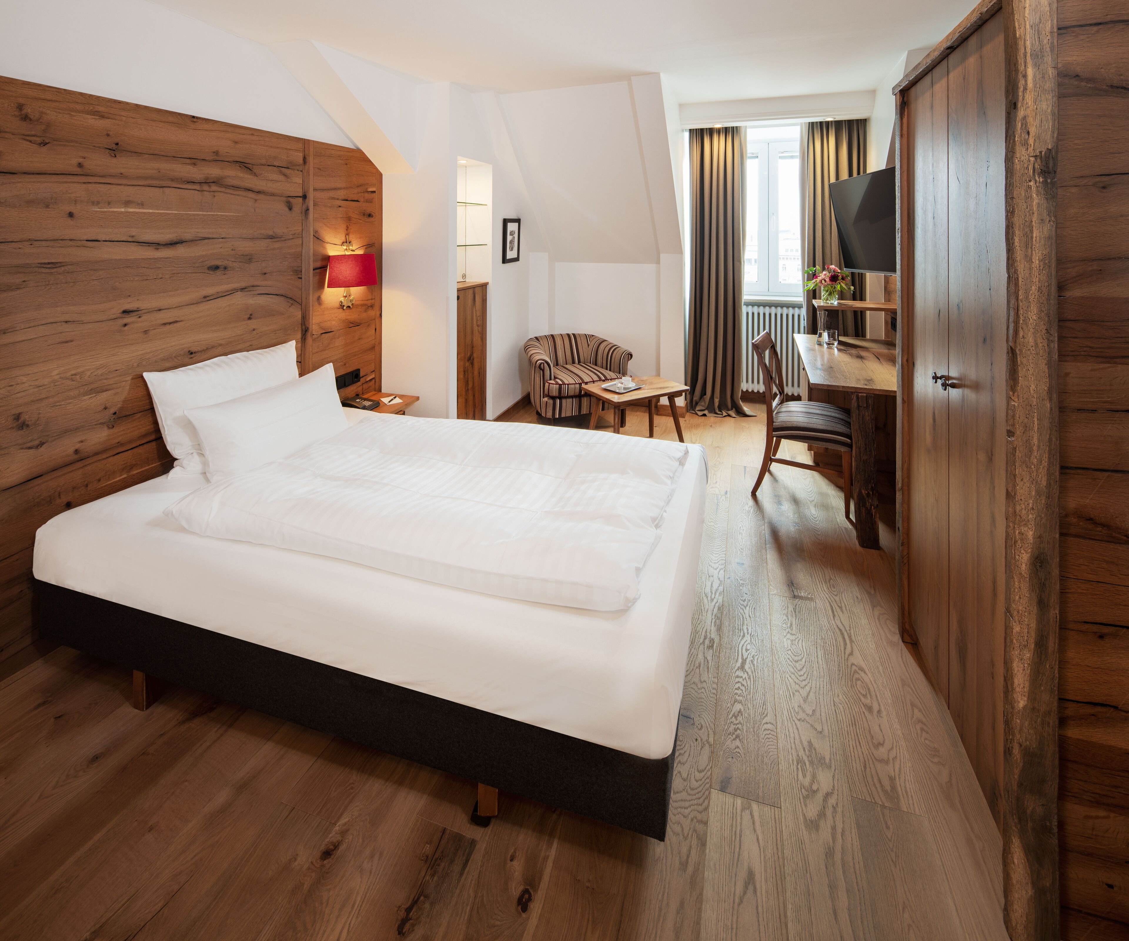 A hotel room with a large bed, wooden headboard, a desk with a chair, a TV, and a view of the city through the window.