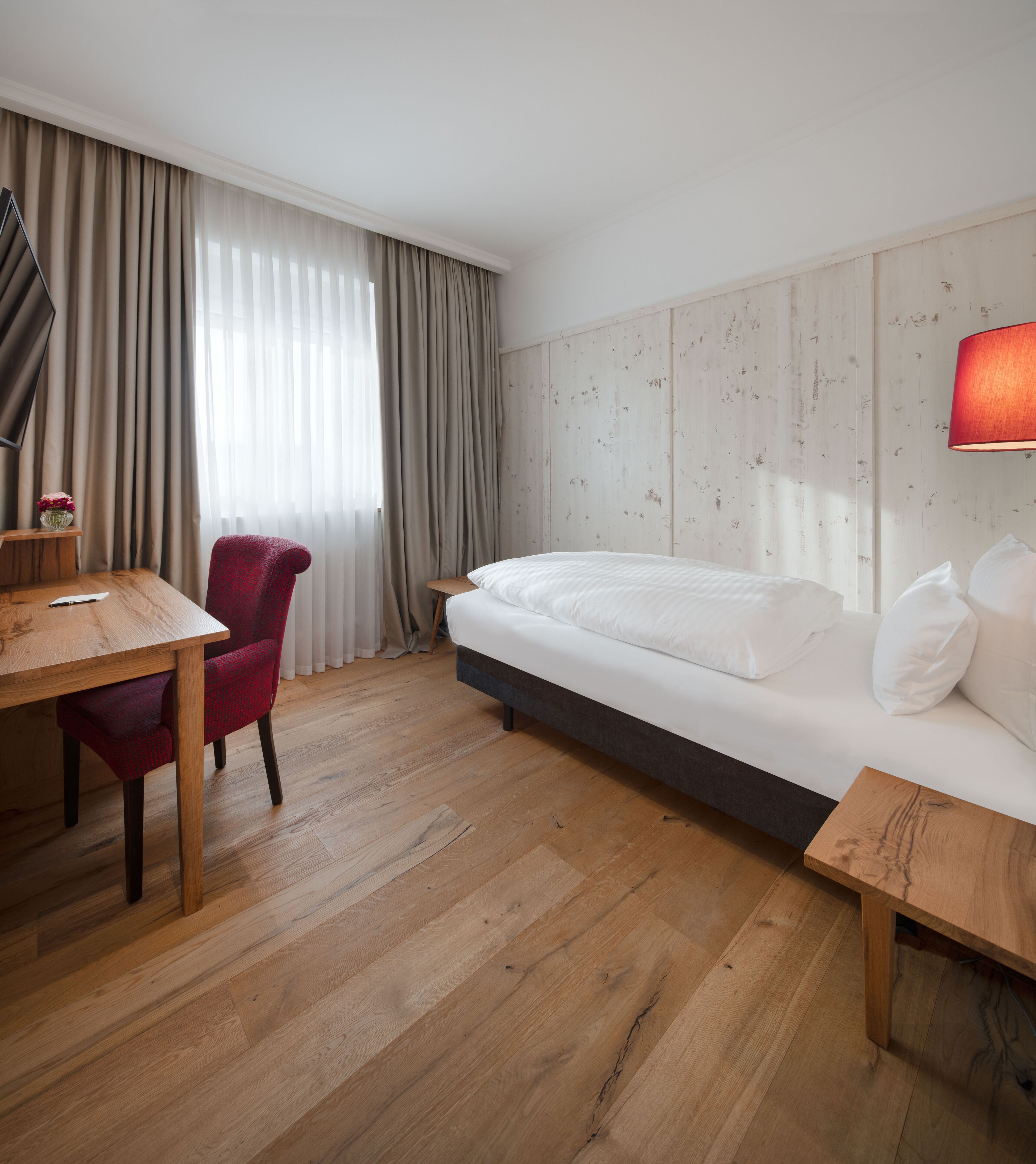 A hotel room with a wooden floor, a bed with white bedding, a red chair, a wooden desk, and a wall-mounted lamp.