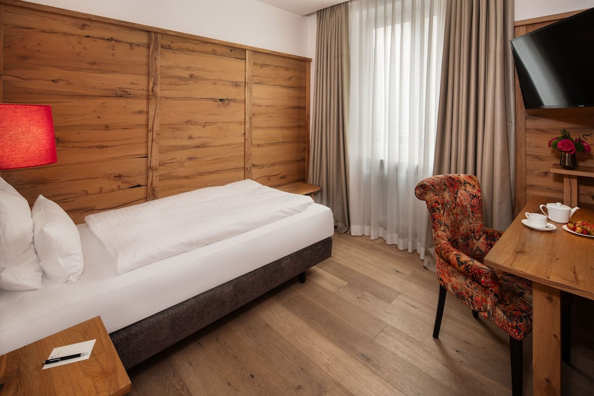 Hotelkamer met een houten hoofdeinde, een bed met witte lakens, een rode lamp, een bloemenstoel, een houten tafel met een theepot en kopjes, en een televisie aan de muur.
