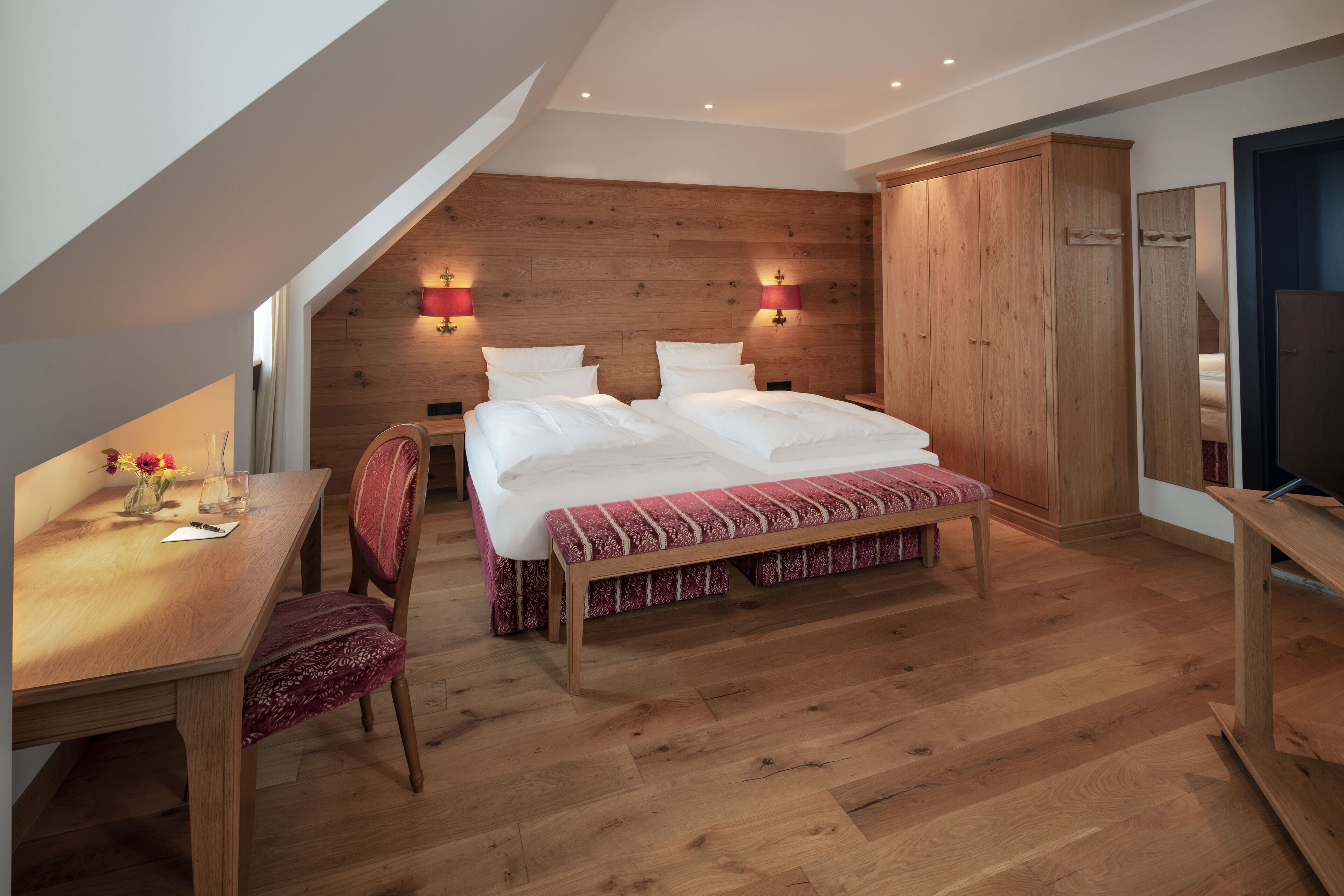 A bedroom with a wooden headboard, a bed with white bedding, a wooden desk with a chair, and a television on a stand.