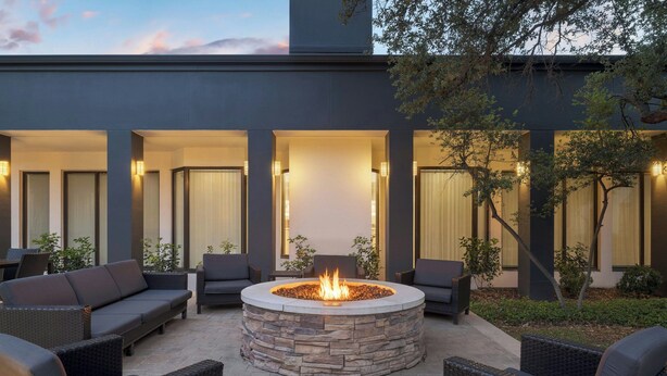Un espace extérieur moderne, aménagé avec un foyer en pierre, des meubles en osier et un bâtiment doté de grandes fenêtres et d’un éclairage chaleureux.