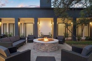 Een modern terras met een vuurkorf van steen, rieten meubilair en een gebouw met grote ramen en warme verlichting.