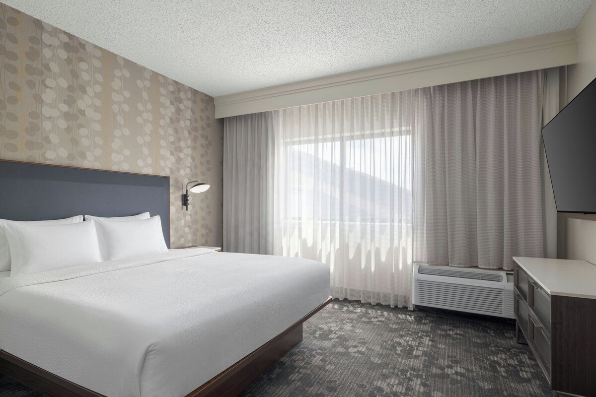 A hotel room with a large bed, a desk, a flat-screen TV, a window with curtains, and a wall with a patterned design.
