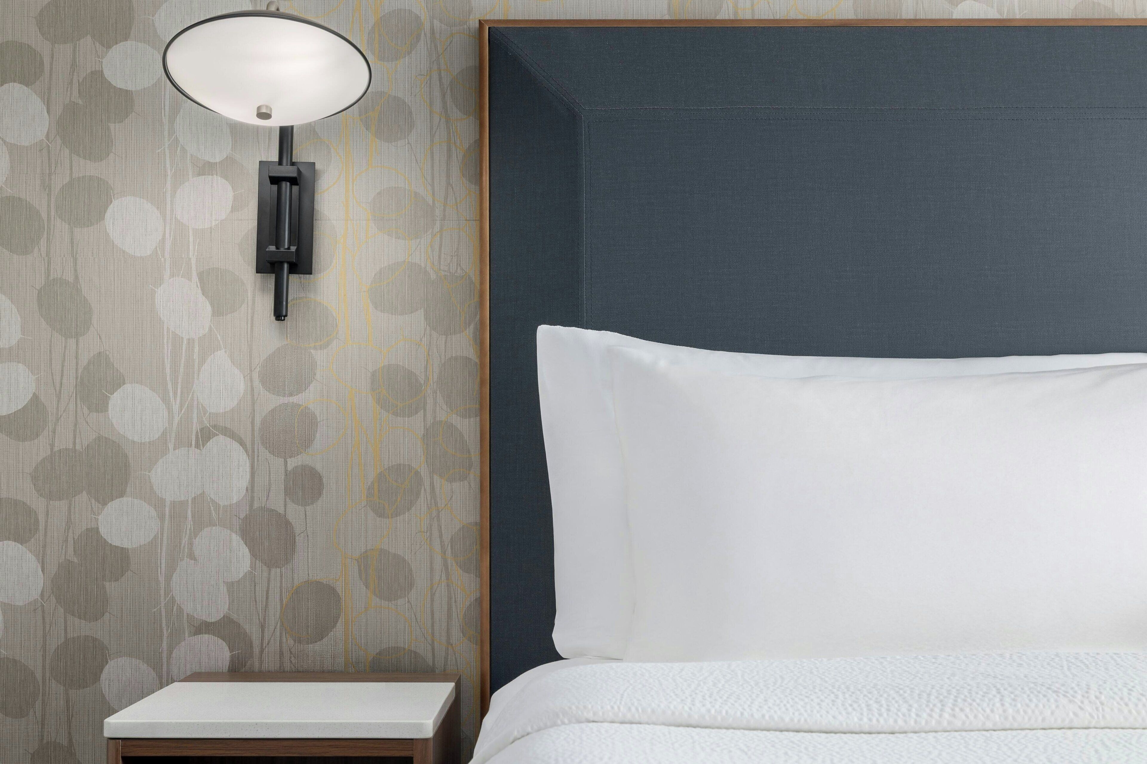 Una cama bien hecha con un almohadón blanco y cabecera, una mesita de noche pequeña y una lámpara de pared.