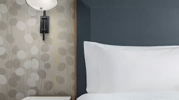 Una cama bien hecha con un almohadón blanco y cabecera, una mesita de noche pequeña y una lámpara de pared.