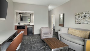 Une chambre d’hôtel moderne dotée d’une kitchenette, d’un canapé gris, d’une table basse en bois et d’un grand tableau abstrait accroché au mur.