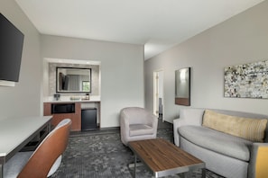 A modern hotel room with a kitchenette, a grey sofa, a wooden coffee table, and a large abstract painting on the wall.