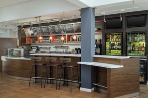 A modern bar area with a wooden counter, bar stools, hanging pendant lights, a menu board, and a refrigerated bar with various bottles. - Courtyard by Marriott Dallas-Fort Worth/Bedford (Bedford)