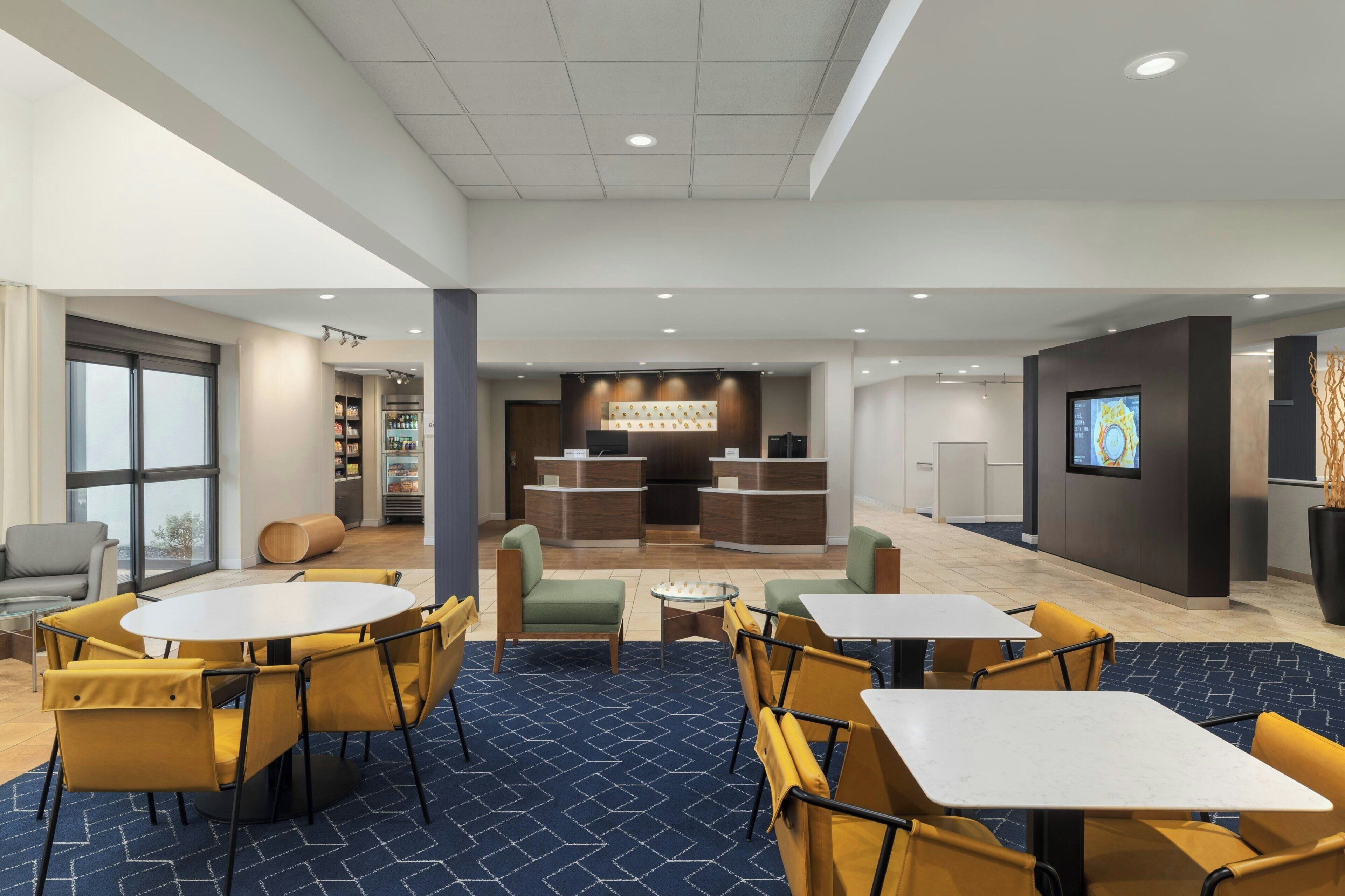A hotel lobby with a reception desk, seating area with tables and chairs, a coffee machine, and a large window.