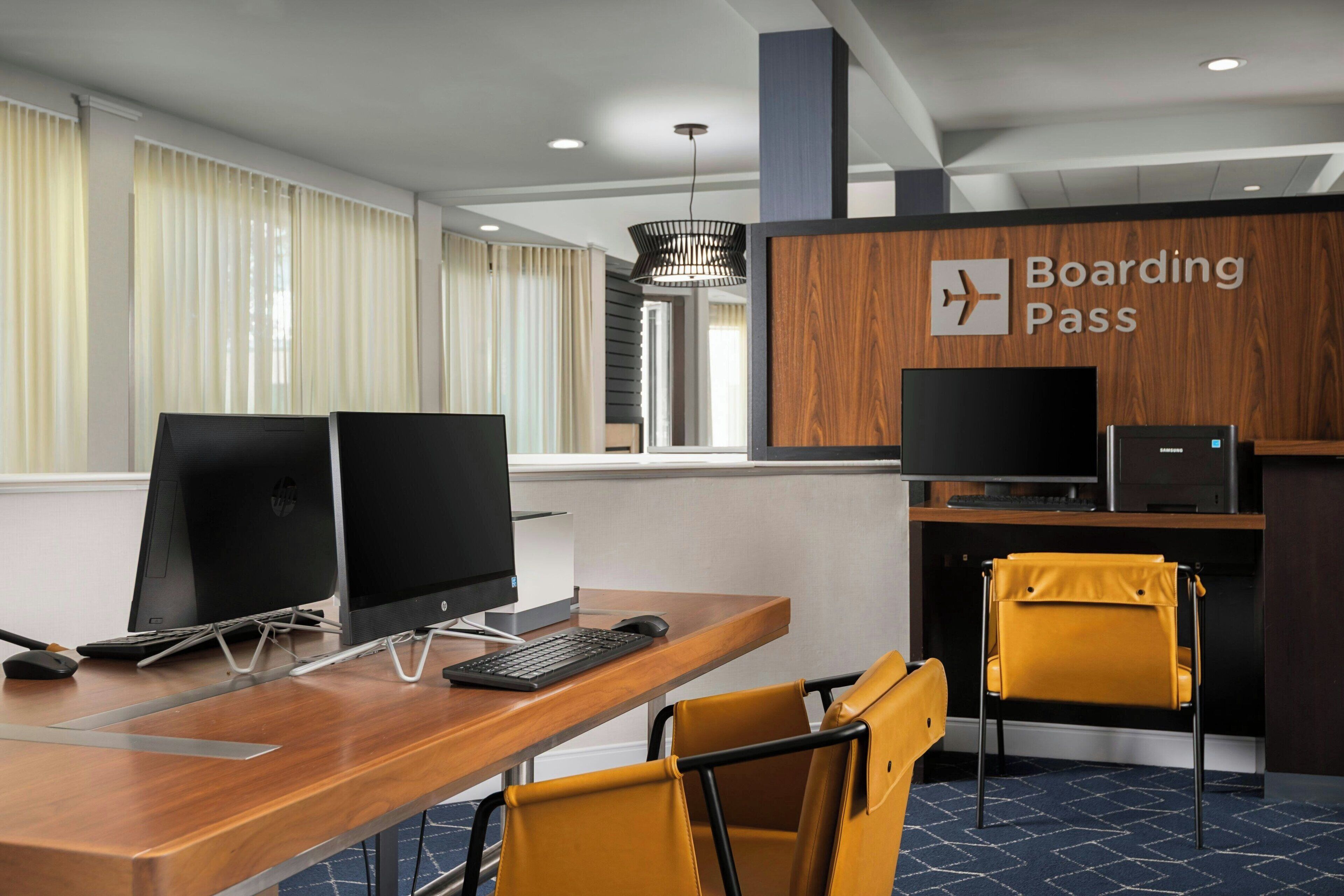 Une chambre d’hôtel moderne avec un bureau en bois, deux écrans, un clavier et une chaise. Un panneau indique l’endroit où se trouve le comptoir pour l’enregistrement des passagers.