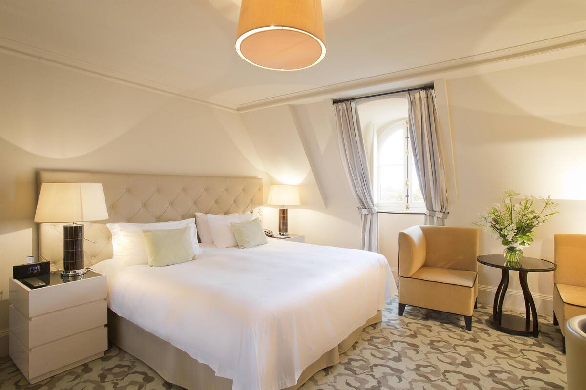 Une chambre d’hôtel avec un grand lit, deux tables de chevet, une chaise, une petite table avec un vase et une fenêtre avec des rideaux.