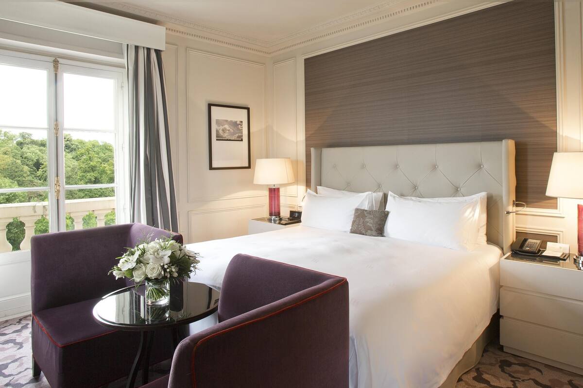 Une chambre d’hôtel avec un grand lit, deux tables de chevet, une chaise, une petite table ornée d’un vase de fleurs et une vue sur la verdure par la fenêtre.