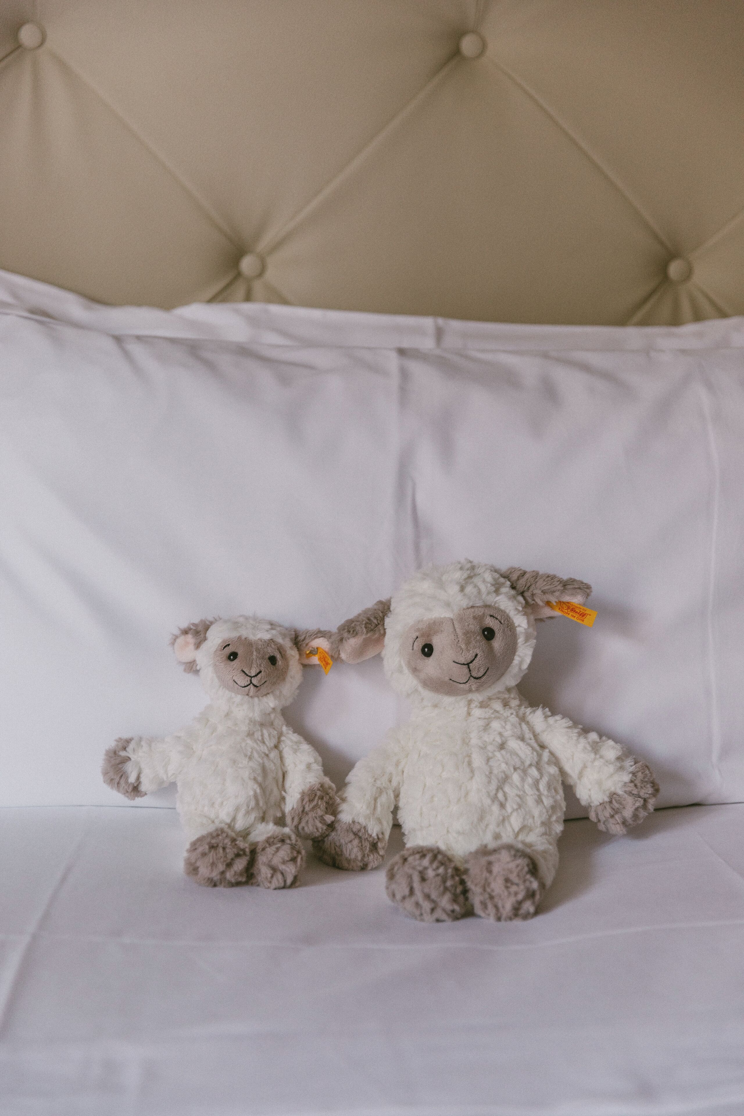 Two plush sheep toys on a white bed with a tufted headboard.