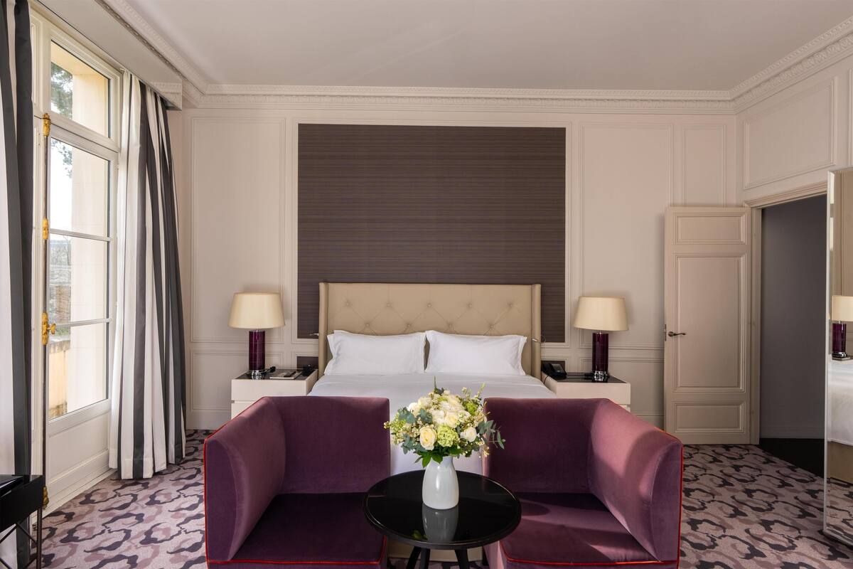 Une chambre d’hôtel avec un grand lit, deux tables de chevet, un canapé violet, une petite table ronde ornée d’un vase de fleurs et une moquette à motifs.