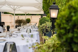 Une rangée de tables rondes, dressées pour un repas en plein air, avec des nappes blanches et des couverts en argent.
