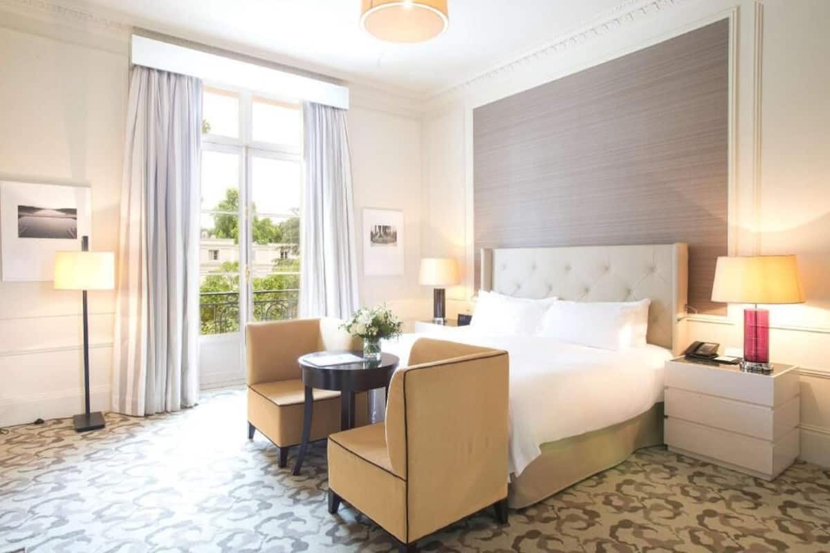 Une chambre d’hôtel avec un grand lit, deux tables de chevet, une table ronde, deux chaises, une fenêtre avec des rideaux et une vue sur la verdure à l’extérieur.