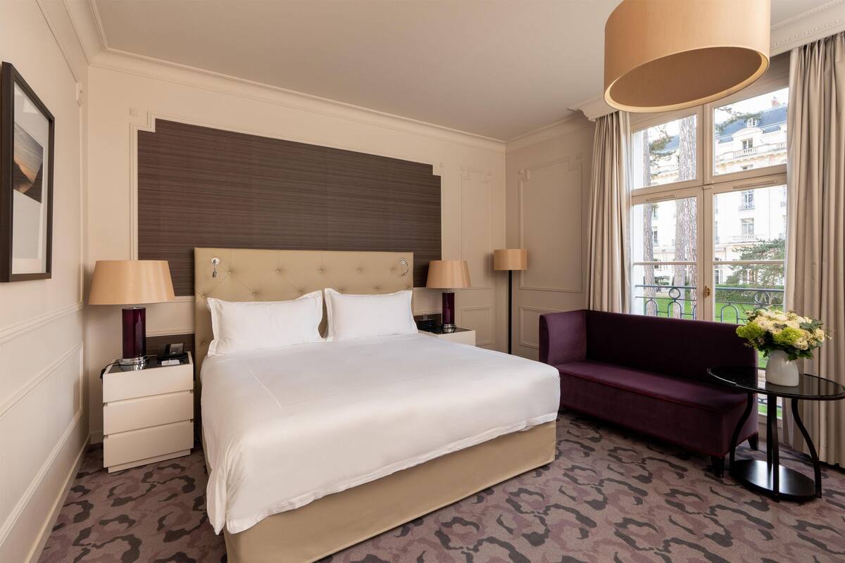 Une chambre d’hôtel avec un grand lit, deux tables de chevet, un canapé, une petite table ornée d’un vase de fleurs et une fenêtre avec des rideaux.