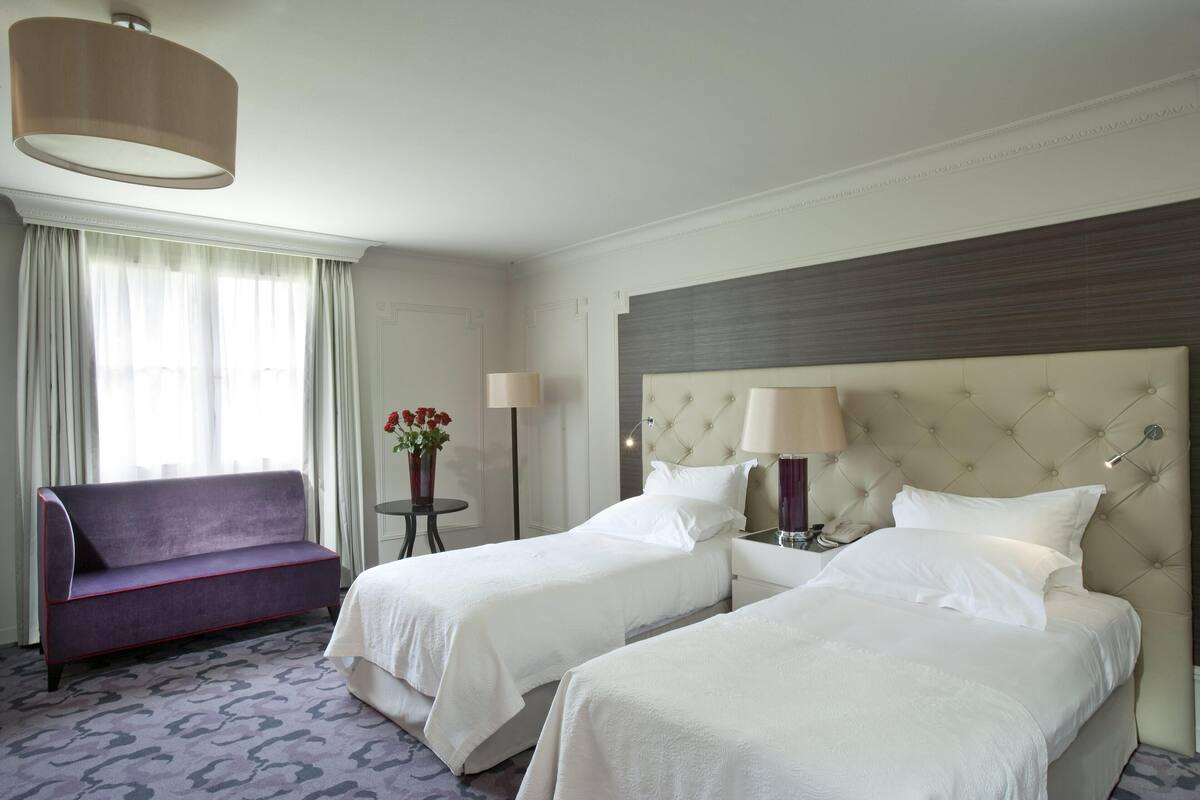 Une chambre d’hôtel avec deux lits, un canapé, une petite table avec une lampe et une grande fenêtre avec des rideaux.