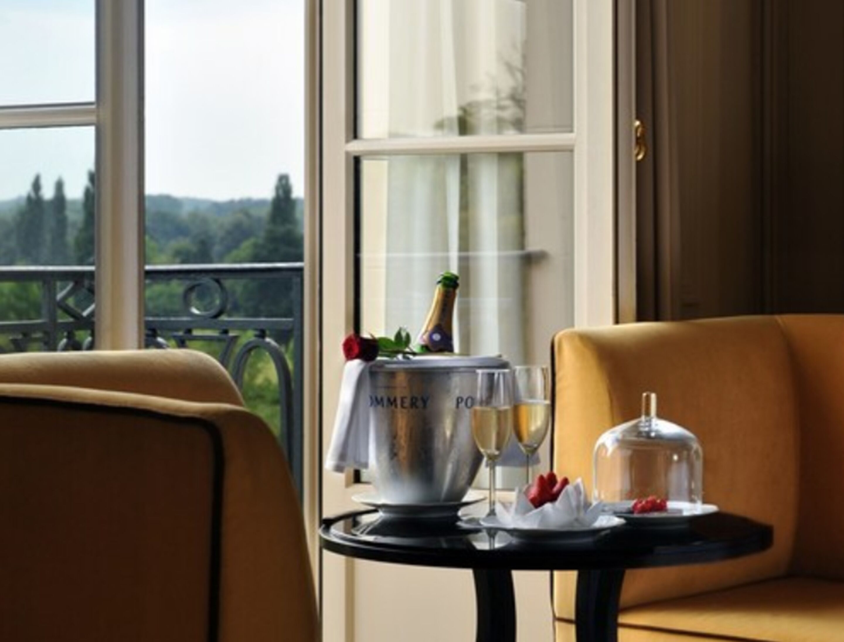 Une table à manger avec une bouteille de champagne, des verres et un plat couvert, le tout placé devant une fenêtre donnant sur un paysage verdoyant.