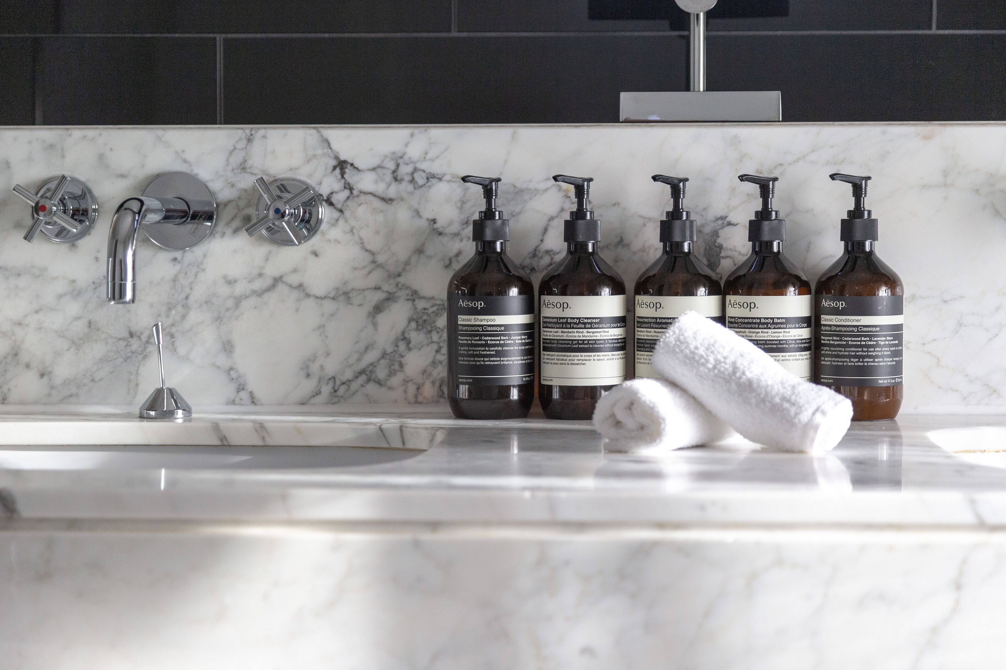 Een badkamer met een marmeren aanrecht, een wastafel en zes flessen Aesop shampoo en conditioner.