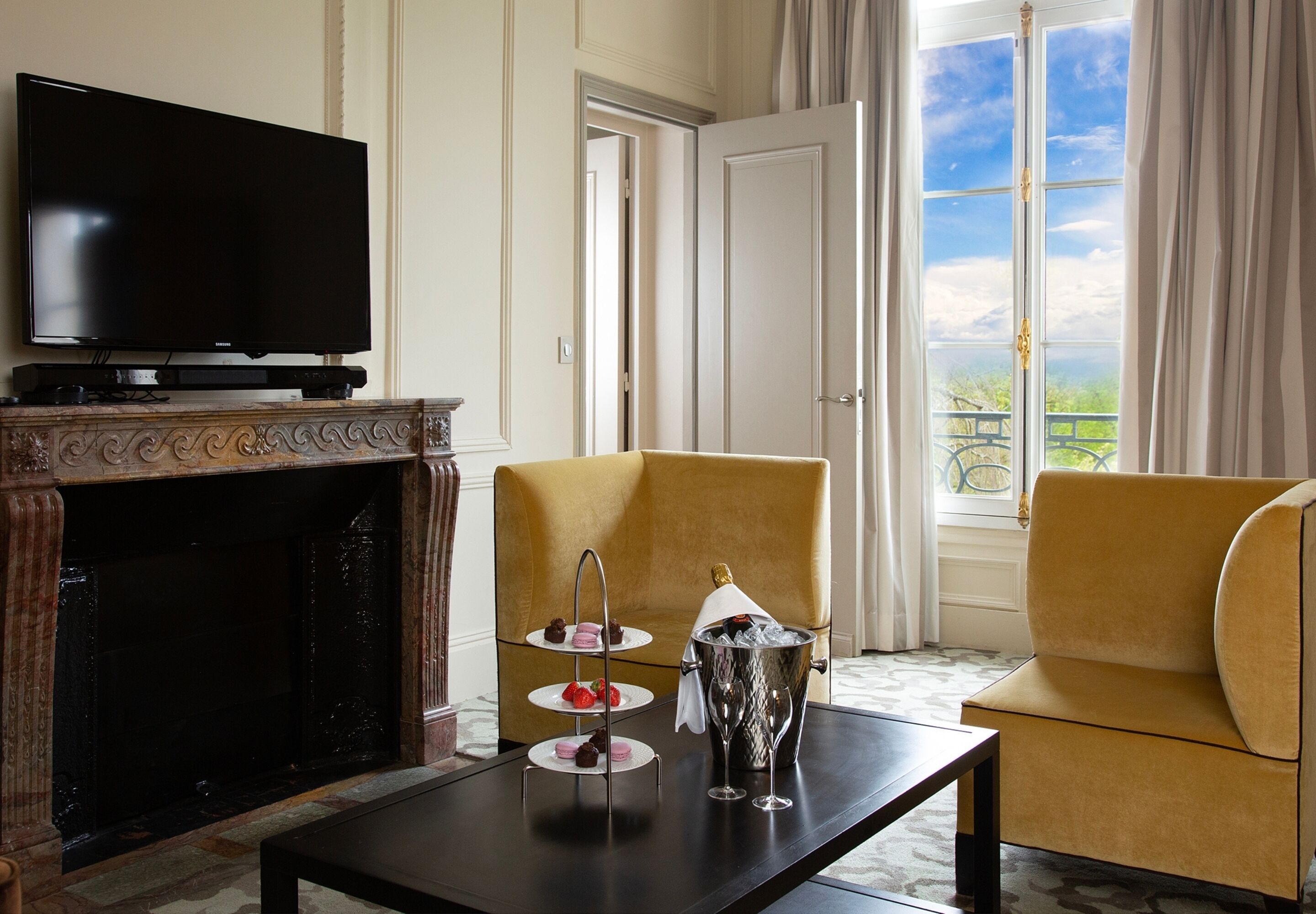 Une pièce dotée d’un téléviseur à écran plat, d’une cheminée, d’un fauteuil jaune, d’une petite table avec une bouteille de champagne et des verres, et offrant une vue sur le balcon.