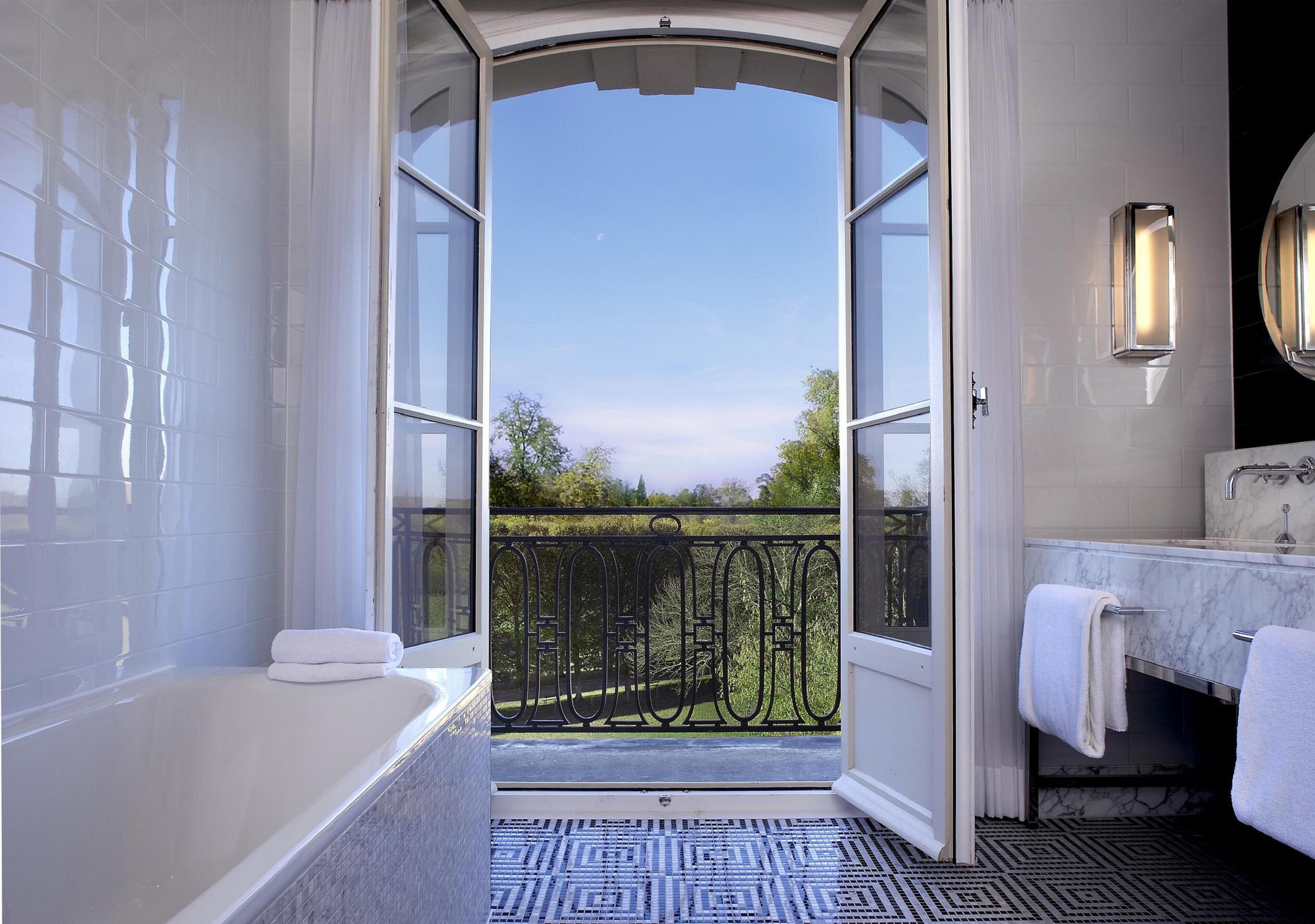 Une salle de bain avec une baignoire, un lavabo et des toilettes. Il y a un balcon avec une rambarde en métal et une vue sur les arbres et un ciel dégagé.