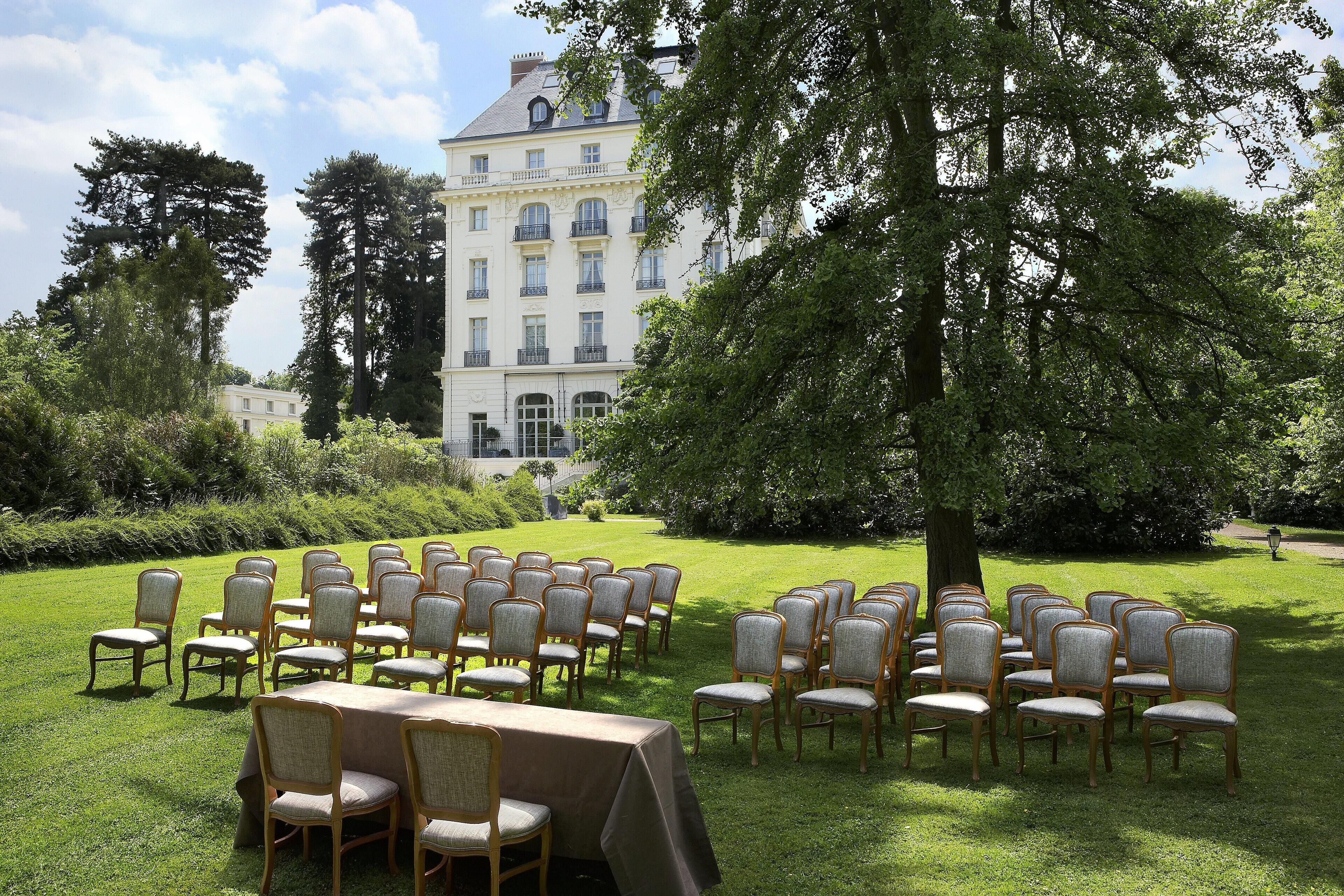 A formal outdoor seating arrangement with chairs and a table, set against a backdrop of a large building and trees.