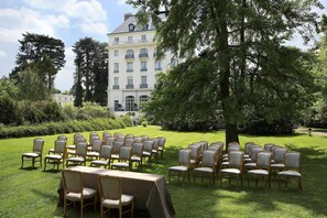 A formal outdoor seating arrangement with chairs and a table, set against a backdrop of a large building and trees.