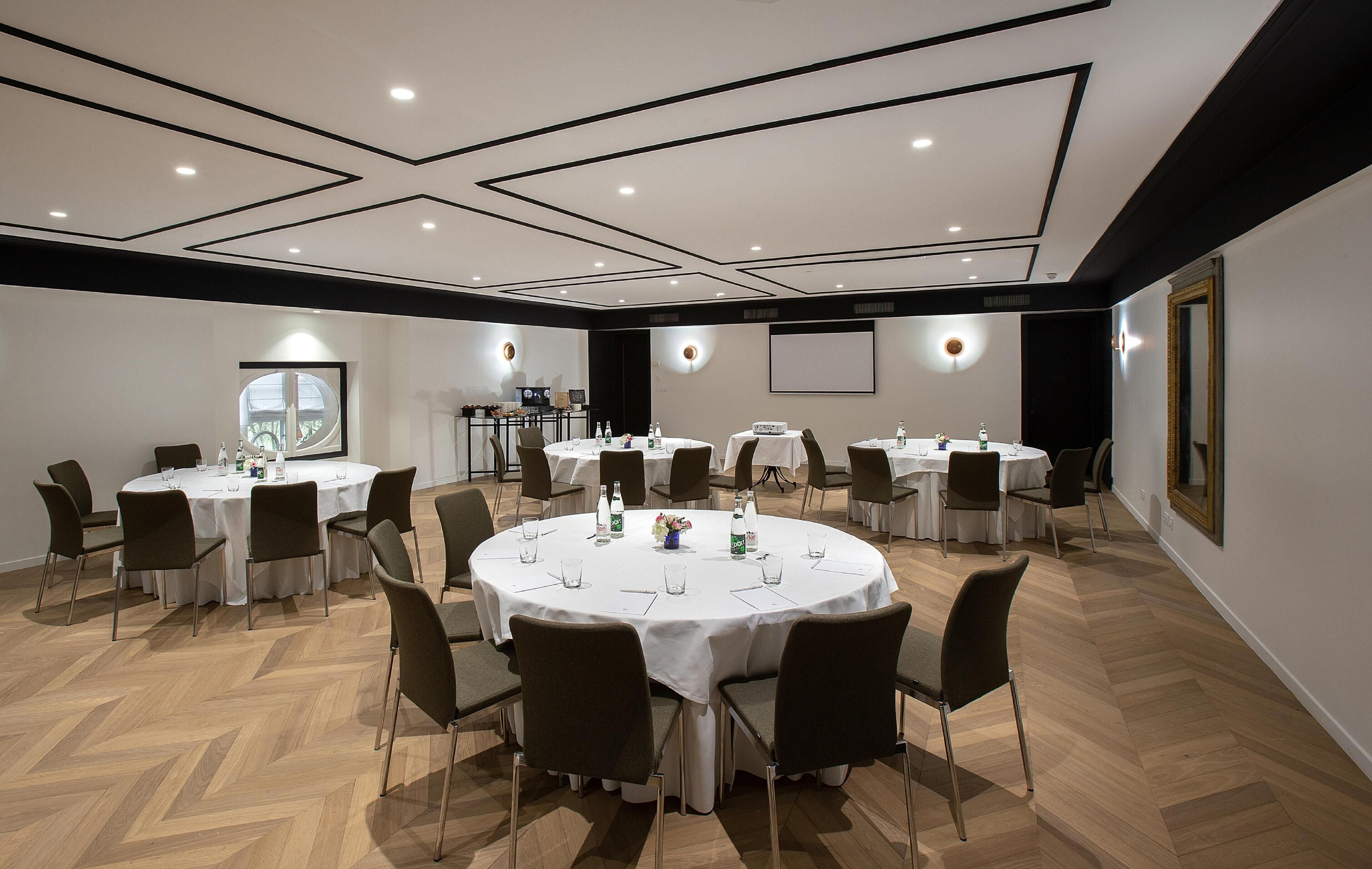 Une salle de conférence dotée de plusieurs tables rondes, chacune entourée de chaises, et équipée de tables, de chaises et d’un écran de projection.