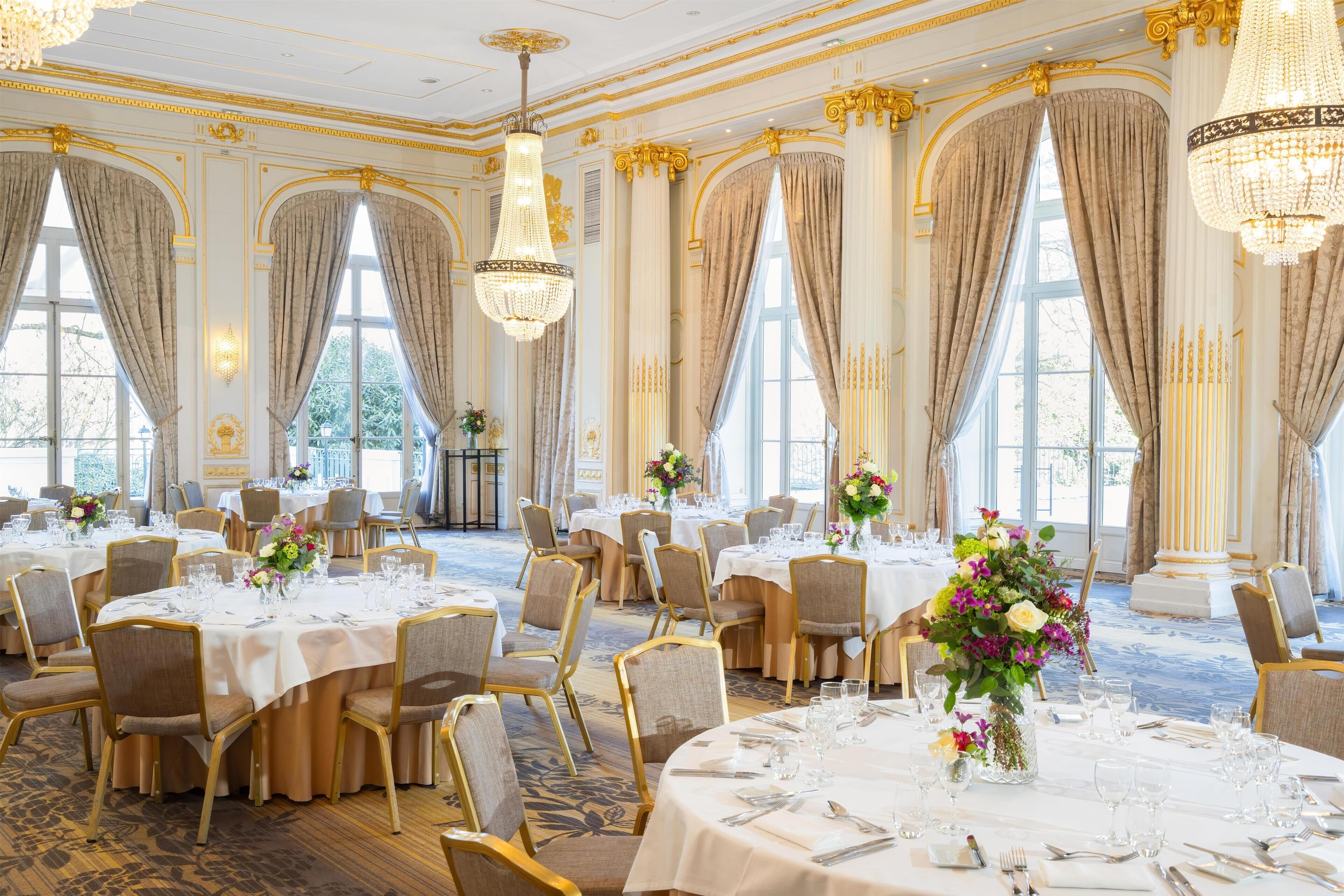 Une salle à manger élégante, avec des tables rondes dressées pour le repas, de grandes fenêtres ornées de rideaux et des lustres.