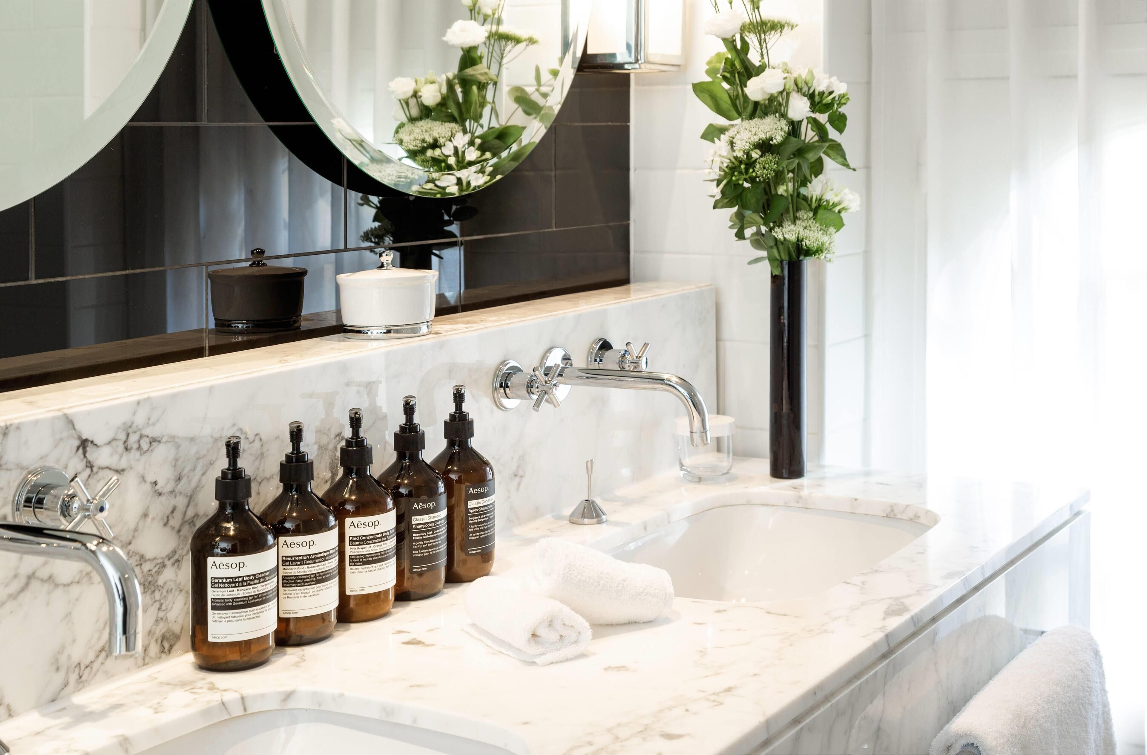 A bathroom with a marble sink, three bottles of Aesop skincare products, a white towel, and a vase with flowers.