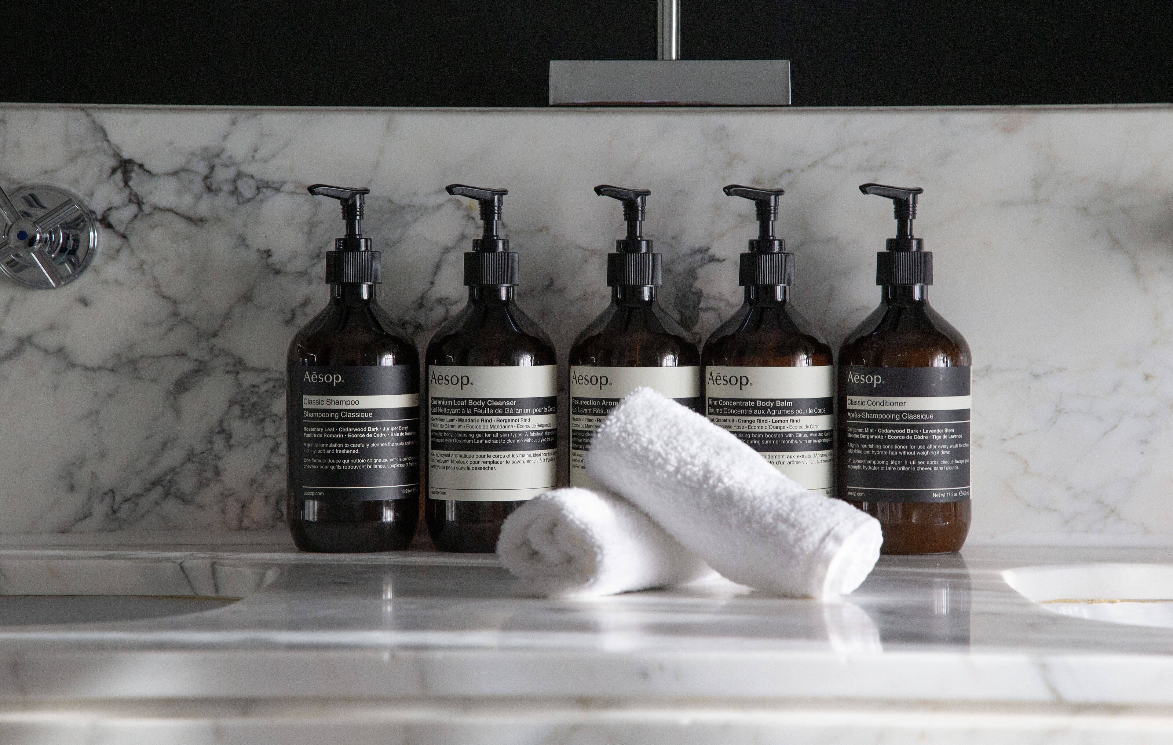Um banheiro com bancada de mármore, cinco frascos de produtos de cuidados com a pele Aesop e uma toalha enrolada.