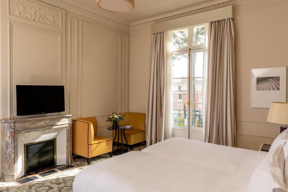 Une chambre d’hôtel avec un grand lit, un téléviseur fixé au-dessus d’une cheminée, une petite table avec un vase de fleurs, un fauteuil jaune et une fenêtre avec des rideaux.