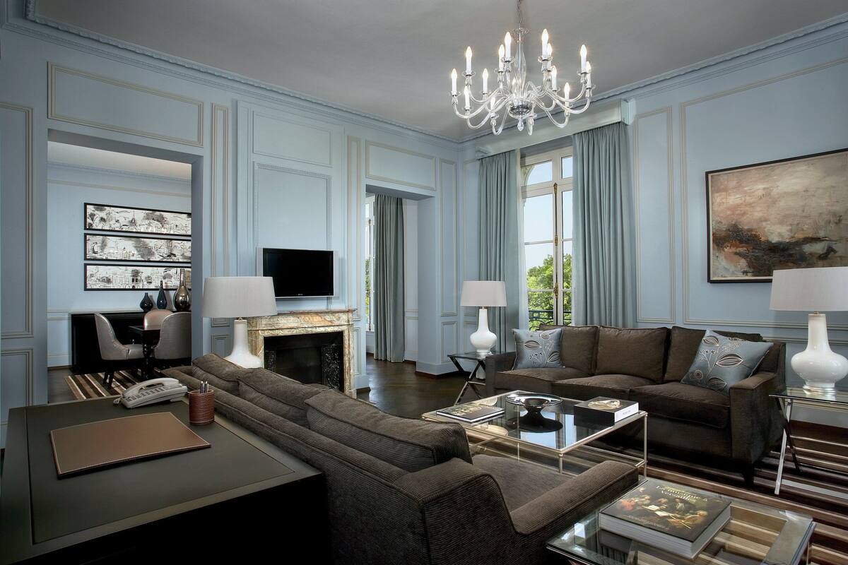 Un salon spacieux comprenant un grand canapé, un canapé plus petit, une table basse et une cheminée. On y trouve deux lampes, un lustre et un téléviseur à écran plat fixé au mur.