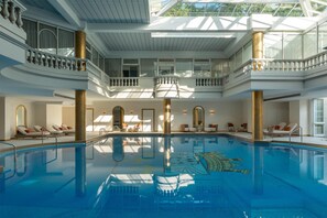 A large indoor swimming pool with a glass ceiling, multiple levels with balconies, and sun loungers around the pool.