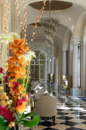 Un vaste hall d’hôtel avec un sol à carreaux, des fauteuils élégants, un lustre et une grande fenêtre.