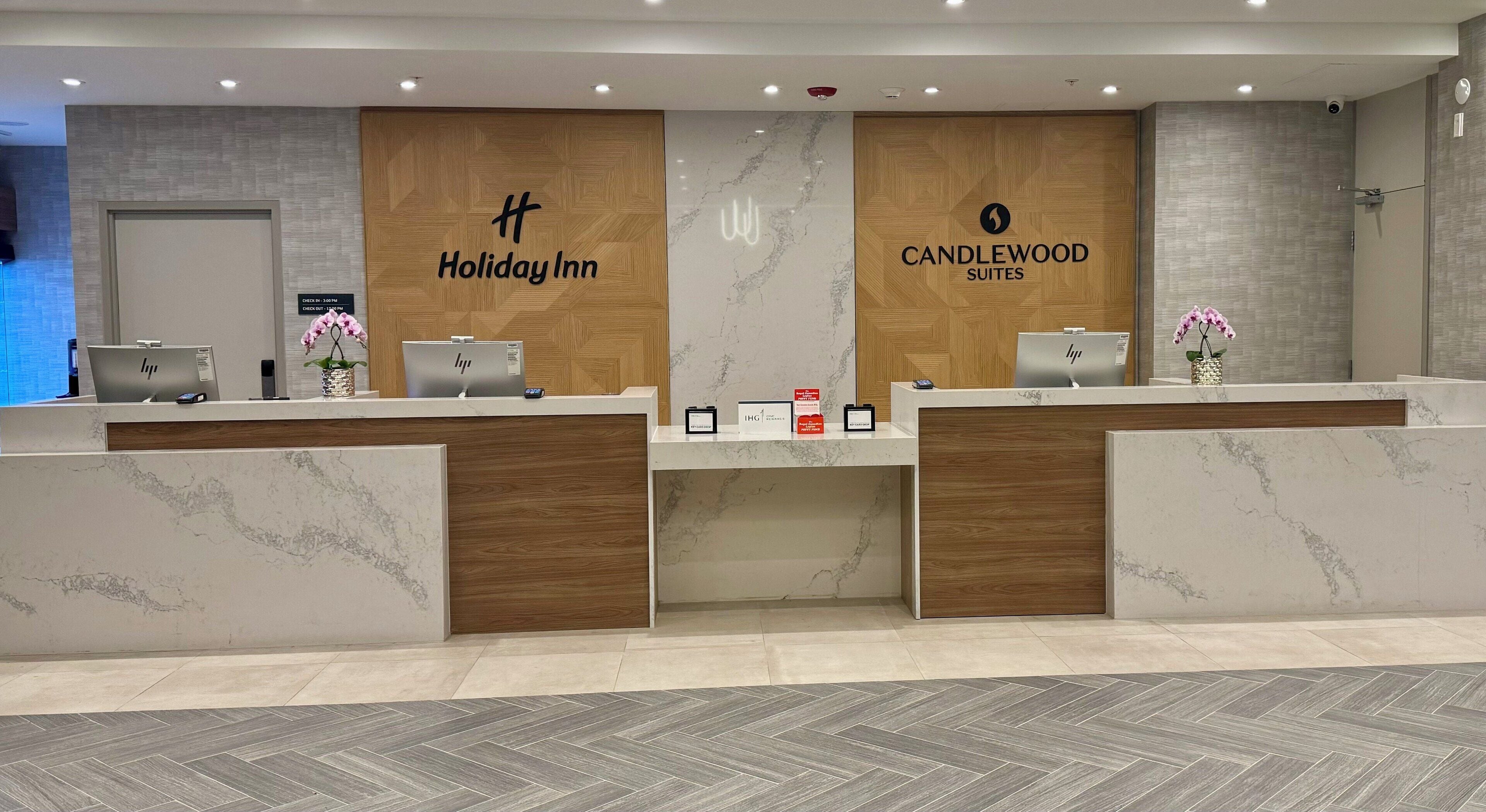 Zona de recepción de hotel con dos mostradores, uno identificado como 'Holiday Inn' y el otro como 'Candlewood Suites'. Hay recepcionistas en cada mostrador, y el suelo presenta un diseño de espiga.